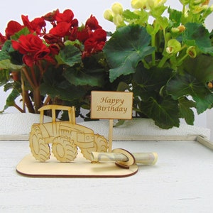 May include: Wooden tractor money holder with a sign that says "Happy Birthday". The tractor is brown and has a green money slot. The holder is on a white surface.