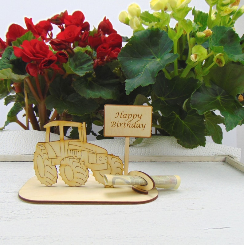 May include: Wooden tractor money holder with a sign that says "Happy Birthday". The tractor is brown and has a green money slot. The holder is on a white surface.