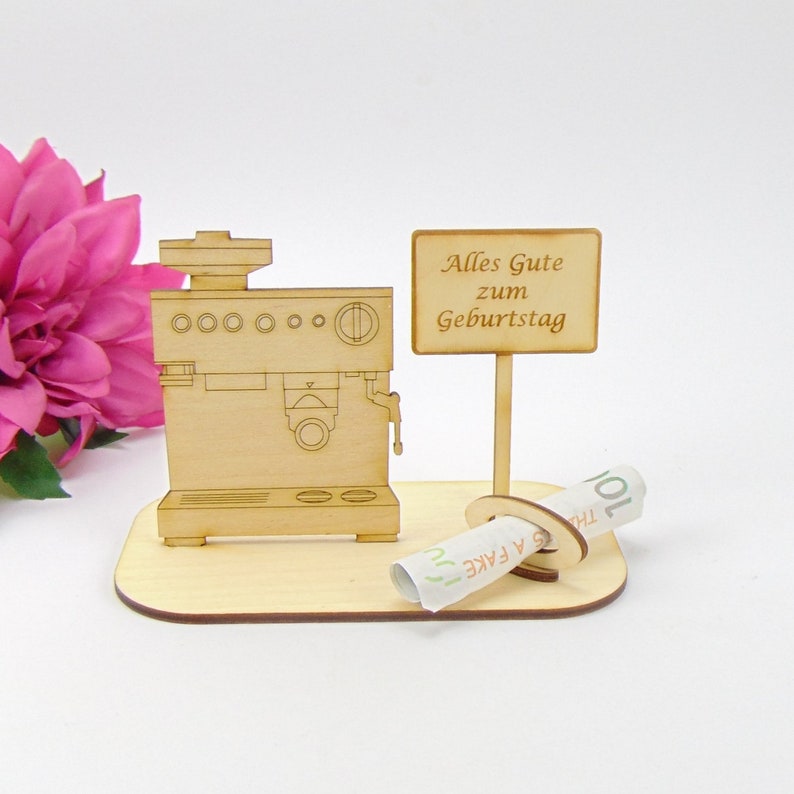 Pu&ograve; includere: Un modello in legno di una macchina per caff&egrave; espresso con un cartello che recita "Alles Gute zum Geburtstag" e un foglio di carta arrotolato con le parole "A FAKE". Il modello &egrave; su una base di legno, con un fiore rosa sullo sfondo.