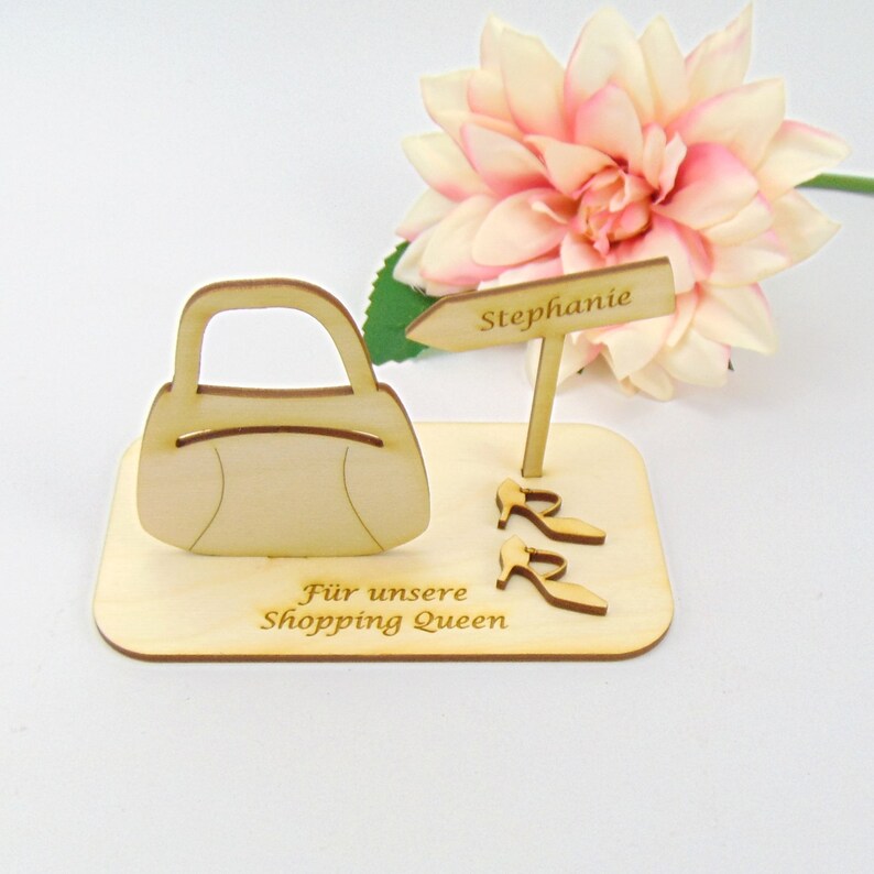 May include: A wooden decorative piece featuring a handbag, two pairs of high heels, and a sign that reads "Stephanie." The base has the text "F&uuml;r unsere Shopping Queen." A pink and white flower is in the background.