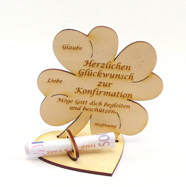 Op de afbeelding: Houten klavertje vier kaart met de tekst "Herzlichen Gl&uuml;ckwunsch zur Konfirmation" en "M&ouml;ge Gott dich begleiten und besch&uuml;tzen." De kaart heeft een gleuf voor een opgerold biljet en een hartvormige basis.