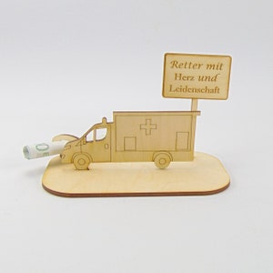 Op de afbeelding: Houten ambulance-figuur met een rood kruis aan de zijkant en een bord met de tekst "Retter mit Herz und Leidenschaft". Een opgerold briefje steekt uit de zijkant van de ambulance.