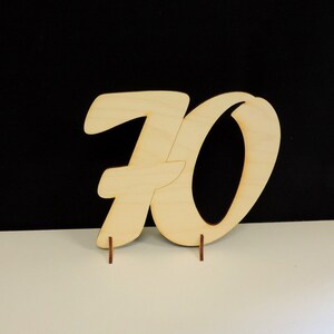 May include: Wooden number 70 in a cursive style, standing on a white surface. The number is made of light brown wood and is approximately 12 inches tall.