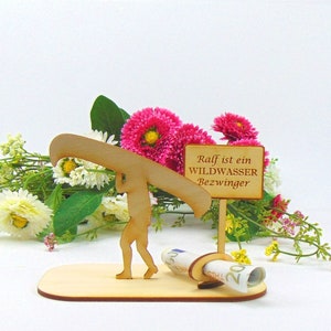 May include: A light-coloured wooden cutout figure carrying a canoe on their shoulders, standing on a flat base. Next to the figure is a small sign with the text "Ralf ist ein WILDWASSER Bezwinger" and a circular holder containing a rolled 20 Euro bill.