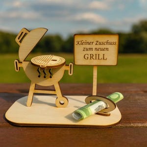 Könnte beinhalten: Eine Holzminiatur eines Grills mit offenem Deckel, einem Schild mit der Aufschrift "Kleiner Zuschuss zum neuen GRILL" und einem aufgerollten Geldschein. Die Miniatur steht auf einem Holzsockel.