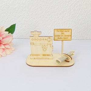 Pu&ograve; includere: Un modello di macchina per caff&egrave; espresso in legno con un cartello che recita "Du bist nicht DU, wenn DU keinen Kaffee hast!". Un oggetto decorativo a tema caff&egrave; con una carta arrotolata e una base in legno.
