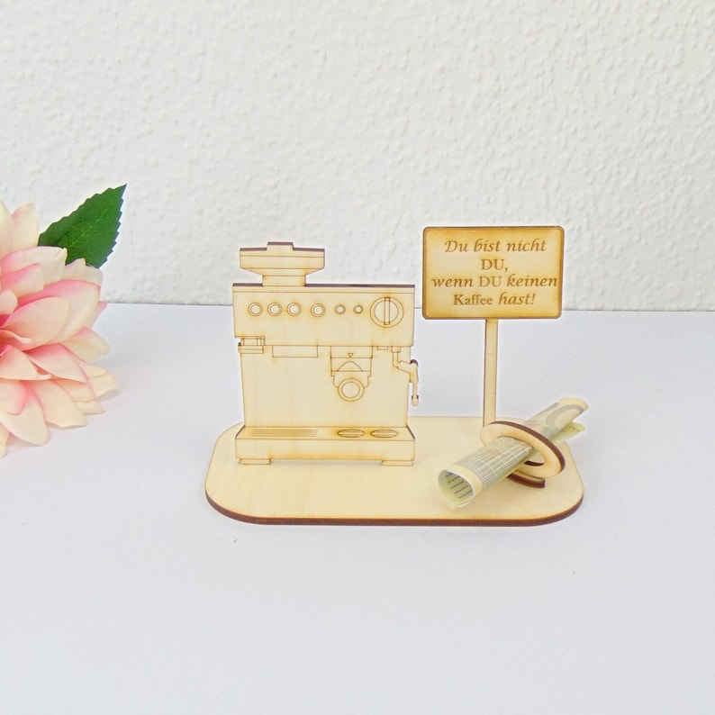 Pu&ograve; includere: Un modello di macchina per caff&egrave; espresso in legno con un cartello che recita "Du bist nicht DU, wenn DU keinen Kaffee hast!". Un oggetto decorativo a tema caff&egrave; con una carta arrotolata e una base in legno.