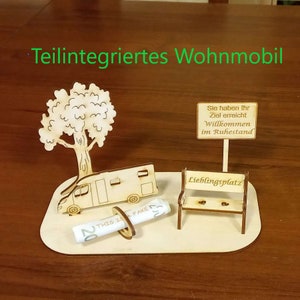 May include: A wooden model of a camper van, a tree, a bench, and a sign that reads "Sie haben Ihr Ziel erreicht Willkommen im Ruhestand" (You have reached your goal Welcome to retirement). The model is on a wooden base and there is a rolled-up fake twenty euro bill next to the camper van.