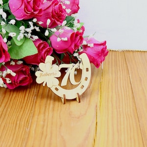 May include: Wooden horseshoe-shaped sign with the number 70 and the name Roberto inscribed on a shamrock. The sign is surrounded by a bouquet of pink roses.