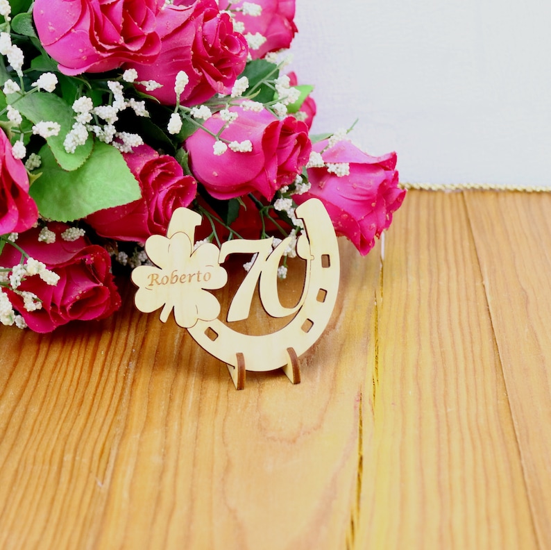 May include: Wooden horseshoe-shaped sign with the number 70 and the name Roberto inscribed on a shamrock. The sign is surrounded by a bouquet of pink roses.