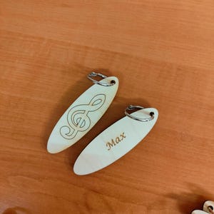 Op de afbeelding: Twee houten sleutelhangers met metalen ringen. De ene sleutelhanger heeft een solsleutel gegraveerd, de andere sleutelhanger heeft de naam "Max" gegraveerd.