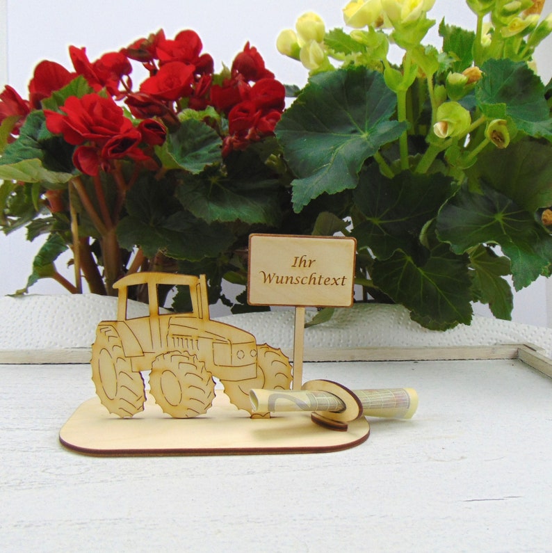 May include: Wooden tractor money holder with a sign that says "Ihr Wunschtext". The tractor is brown and has a slot for money in the back. The sign is attached to a stick that is stuck in the ground.