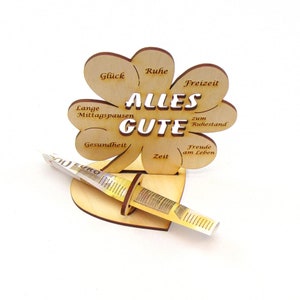May include: A wooden clover-shaped money holder with the words "Alles Gute" in the center. The clover has different wishes written on each leaf, such as "Gl&uuml;ck", "Ruhe", "Freizeit", "Lange Mittagspausen", "Gesundheit", "Zeit", "Freude am Leben", and "zum Ruhestand". A euro bill is inserted into the holder.