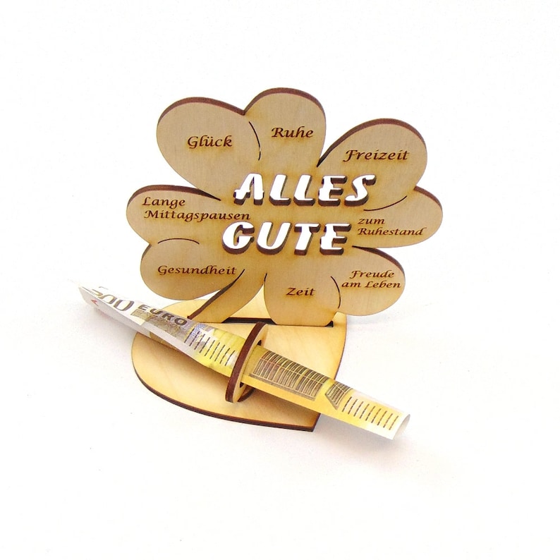 May include: A wooden clover-shaped money holder with the words "Alles Gute" in the center. The clover has different wishes written on each leaf, such as "Gl&uuml;ck", "Ruhe", "Freizeit", "Lange Mittagspausen", "Gesundheit", "Zeit", "Freude am Leben", and "zum Ruhestand". A euro bill is inserted into the holder.