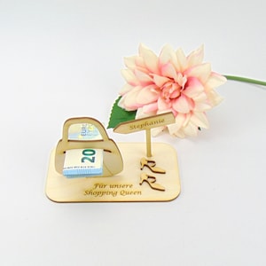 May include: A light-colored wooden display stand featuring a cutout handbag holding a twenty-euro bill, a signpost labeled Stephanie, and two high-heeled shoe cutouts. The base is engraved with the text F&uuml;r unsere Shopping Queen.