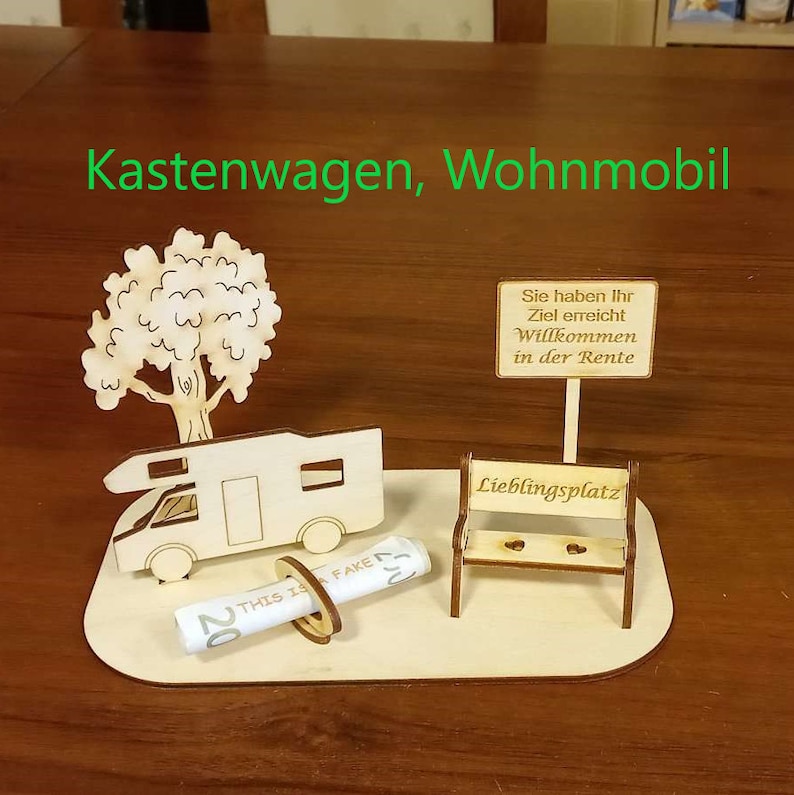 May include: A wooden model of a camper van, a tree, a bench, and a sign that reads "Sie haben Ihr Ziel erreicht Willkommen in der Rente" (You have reached your goal Welcome to retirement). The model is on a wooden base.