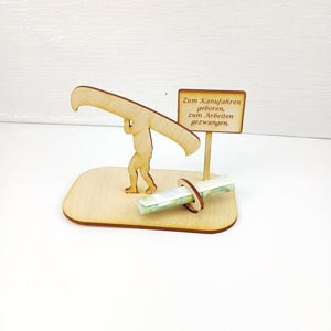 May include: A light-coloured wooden cutout featuring a figure carrying a canoe on their shoulders, standing on a base. A small sign attached to the base displays the text: Zum Kanufahren geboren, zum Arbeiten gezwungen. A rolled banknote is held in a ring.