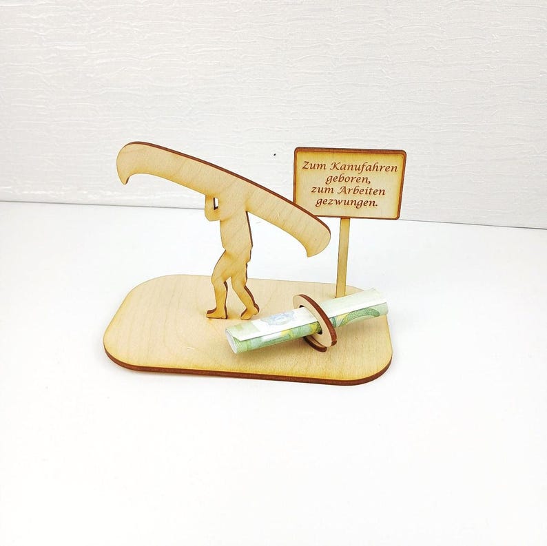 May include: A light-coloured wooden cutout featuring a figure carrying a canoe on their shoulders, standing on a base. A small sign attached to the base displays the text: Zum Kanufahren geboren, zum Arbeiten gezwungen. A rolled banknote is held in a ring.