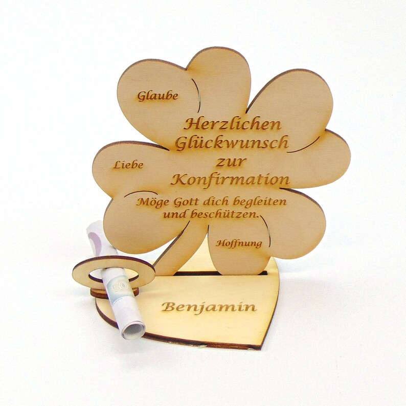 Op de afbeelding: Houten klavertje vier-kaarthouder met de tekst "Herzlichen Gl&uuml;ckwunsch zur Konfirmation" en "M&ouml;ge Gott dich begleiten und besch&uuml;tzen." De kaarthouder heeft een gleuf voor een opgerold biljet en een hartvormige basis met de naam "Benjamin" erop gegraveerd.
