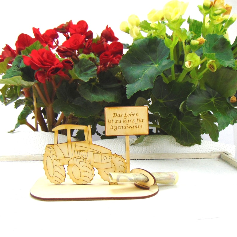 May include: Wooden tractor money holder with a sign that reads "Das Leben ist zu kurz f&uuml;r irgendwann!"  The tractor is brown and the sign is white with black lettering. The money holder is sitting on a wooden base.
