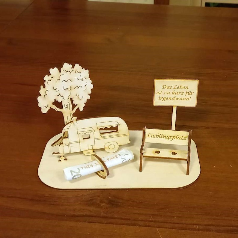 May include: A wooden model of a camper van, a tree, a bench, and a sign that reads "Das Leben ist zu kurz f&uuml;r irgendwann!"  The bench has the words "Lieblingsplatz" on it. A rolled-up piece of paper with the words "THIS IS FAKE" and "20" is on the ground.