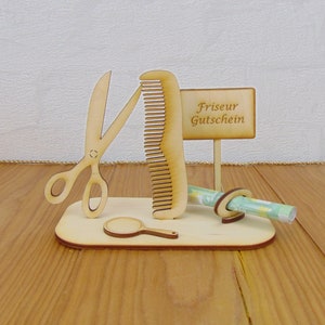 Puede incluir: Soporte de madera con un peine, tijeras y un peque&ntilde;o espejo. Un vale de regalo enrollado se mantiene en su lugar mediante un anillo de madera. El letrero dice "Friseur Gutschein".