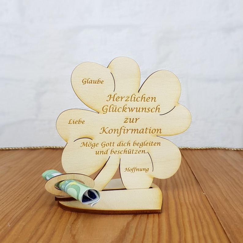 Op de afbeelding: Houten klavertje vier kaart met de woorden "Herzlichen Gl&uuml;ckwunsch zur Konfirmation" en "M&ouml;ge Gott dich begleiten und besch&uuml;tzen." De kaart heeft een gleuf voor geld en de woorden "Glaube", "Liebe", "Hoffnung" staan op de bladeren.