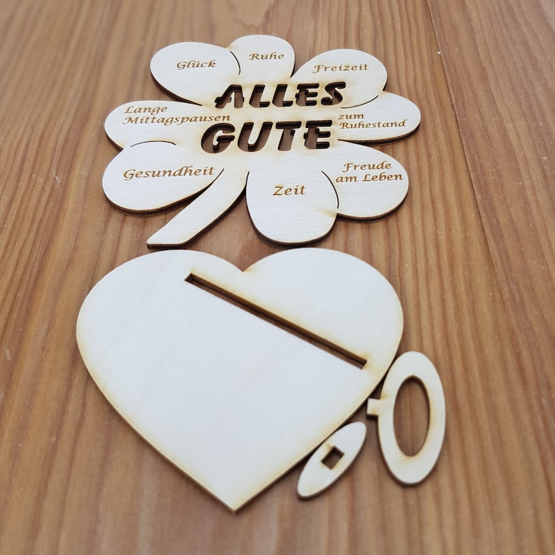 May include: A wooden clover-shaped cutout with the words "ALLES GUTE" in the center. The clover has eight points, each with a different word or phrase written on it, including "Gl&uuml;ck", "Ruhe", "Freizeit", "Lange Mittagspausen", "Gesundheit", "Zeit", "zum Ruhestand", and "Freude am Leben". The clover is accompanied by a wooden heart and a wooden ring.