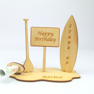 May include: Wooden stand up paddle board birthday card with a sign that says "Happy Birthday" and a slot for money. The base of the card is personalized with the name "B&auml;rbel".