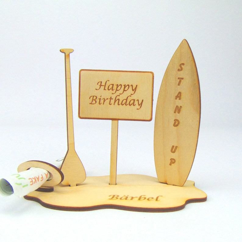 May include: Wooden stand up paddle board birthday card with a sign that says "Happy Birthday" and a slot for money. The base of the card is personalized with the name "B&auml;rbel".