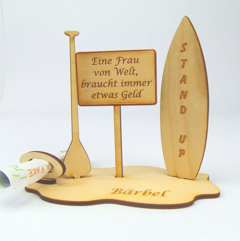 May include: A wooden stand up paddleboard with a sign that reads "Stand Up". There is a wooden paddle and a sign that reads "Eine Frau von Welt, braucht immer etwas Geld". The base of the stand is shaped like a wave and says "B&auml;rbel".