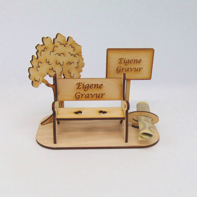 K&ouml;nnte beinhalten: Eine Holzbank mit einem Schild, auf dem "Eigene Gravur" steht, und einem kleinen Baum. Die Bank steht auf einem Holzsockel. Neben der Bank liegt ein zusammengerolltes Blatt Papier auf dem Boden.