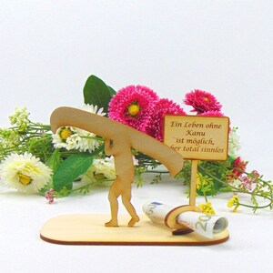 May include: A light-coloured wooden cutout figure carrying a canoe on their shoulders, standing on a flat base. A small sign with the text "Ein Leben ohne Kanu ist m&ouml;glich, aber total sinnlos" is attached, featuring a holder for a rolled banknote.