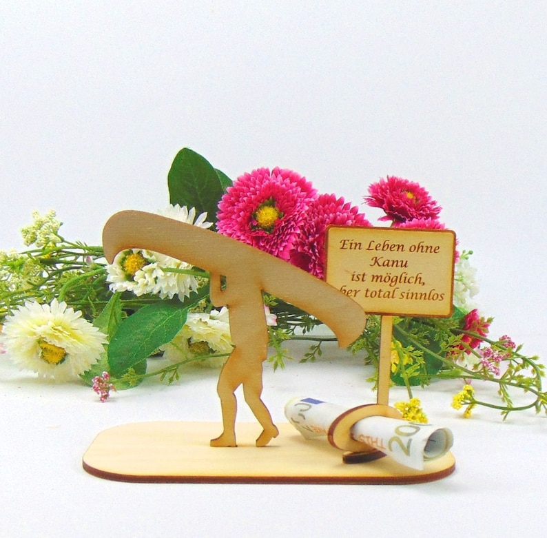 May include: A light-coloured wooden cutout figure carrying a canoe on their shoulders, standing on a flat base. A small sign with the text "Ein Leben ohne Kanu ist m&ouml;glich, aber total sinnlos" is attached, featuring a holder for a rolled banknote.