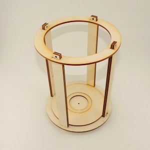 Op de afbeelding: Een onafgewerkt cilindrisch frame van lichtbruin hout met vier verticale steunen en een ronde boven- en onderkant. Het frame is gemaakt van lichtbruin hout en heeft een natuurlijke houtkleur.