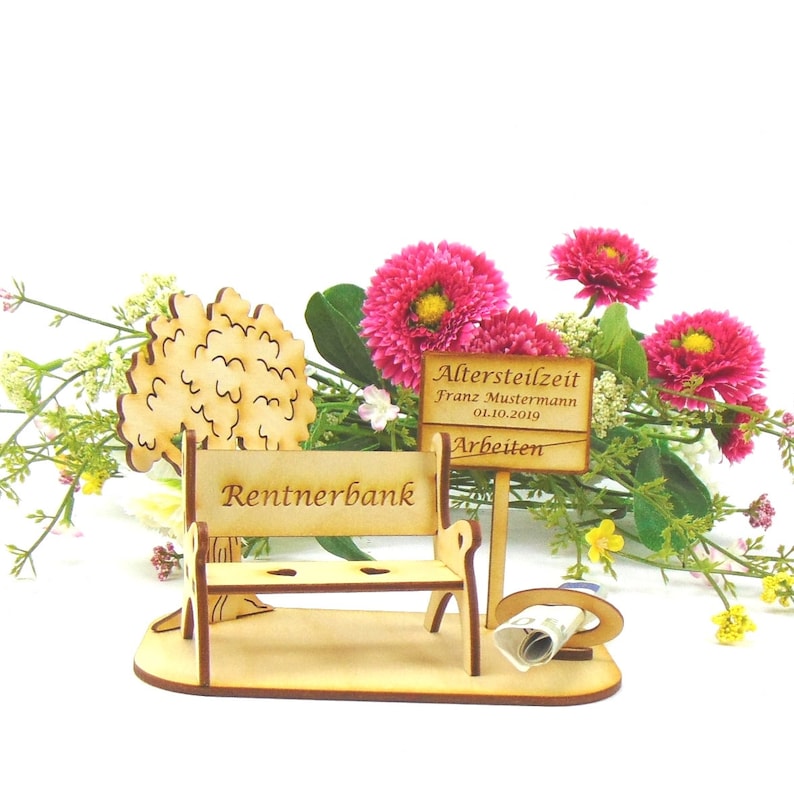 K&ouml;nnte beinhalten: Holzbank mit der Aufschrift "Rentnerbank" und einem Schild mit der Aufschrift "Altersteilzeit Franz Mustermann 01.10.2019 Arbeiten". Die Bank ist von rosa Blumen umgeben.