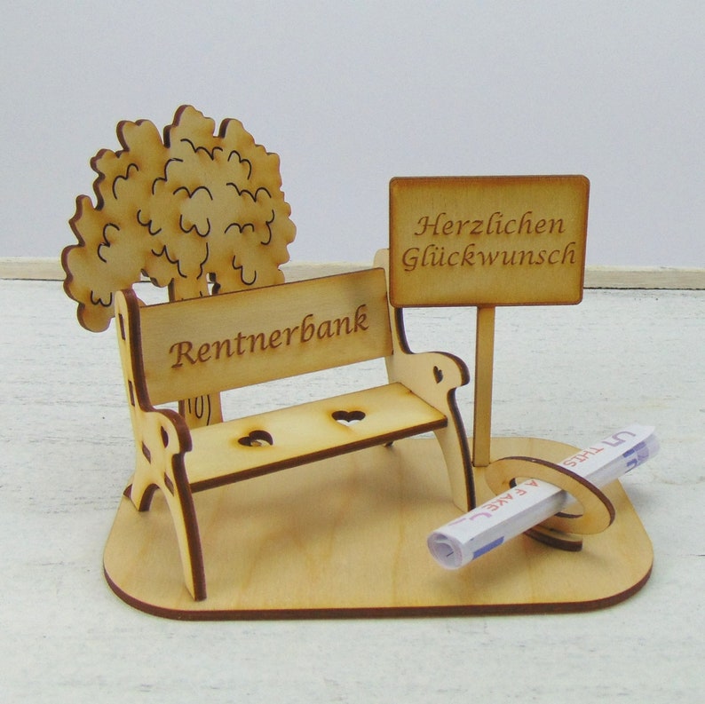K&ouml;nnte beinhalten: Holzbank f&uuml;r den Ruhestand mit einem Baum und einem Schild mit der Aufschrift "Herzlichen Gl&uuml;ckwunsch". Ein zusammengerollter Geldschein liegt auf der Bank.