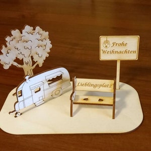 May include: A wooden cutout of a camper, a tree, a bench, and a sign that says "Frohe Weihnachten" (Merry Christmas in German). The bench has the words "Lieblingsplatz" (favorite place in German) on it.