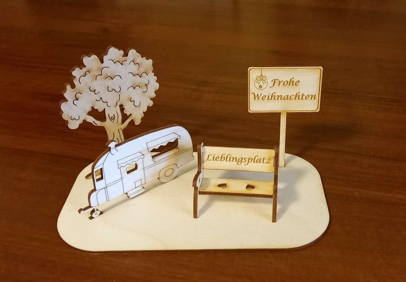 May include: A wooden cutout of a camper, a tree, a bench, and a sign that says "Frohe Weihnachten" (Merry Christmas in German). The bench has the words "Lieblingsplatz" (favorite place in German) on it.