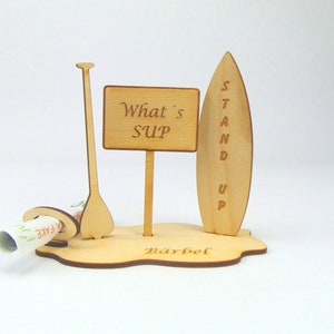 May include: Wooden stand up paddle board (SUP) themed money box with a sign that says "What's SUP" and a paddle. The base of the box is engraved with the name "B&auml;rbel".