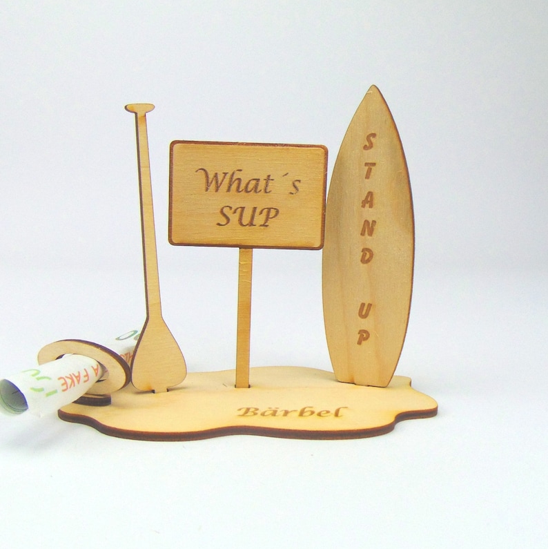 May include: Wooden stand up paddle board (SUP) themed money box with a sign that says "What's SUP" and a paddle. The base of the box is engraved with the name "B&auml;rbel".