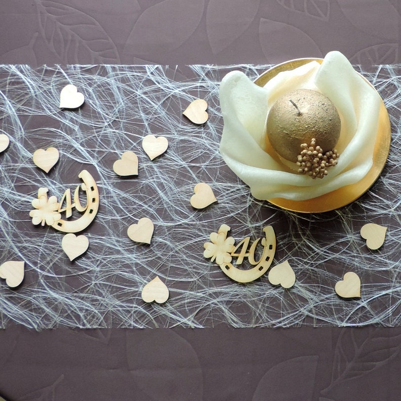 May include: A white table runner with a gold candle in the center. The runner is covered in small wooden hearts and two wooden decorations that say "40" with a horseshoe and a flower.