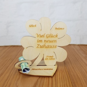 Könnte beinhalten: Ein hölzernes Kleeblatt-Schild mit dem Text "Viel Glück im neuen Zuhause" und den Worten "Glück", "Balance" und "Freude am Leben" auf den Blättern. Das Schild hat einen Schlitz für Geld und eine herzförmige Basis mit dem Text "Mustername".