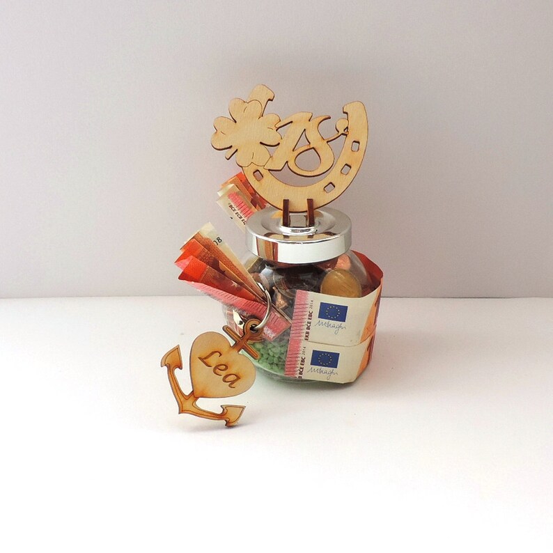 May include: A glass jar filled with money and other small items, decorated with a wooden horseshoe, clover, and the number 18. A wooden heart with the name "Lea" is attached to a wooden anchor.