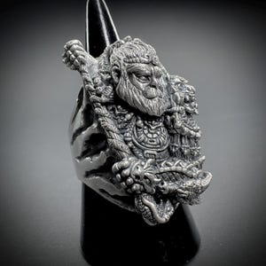 May include: A silver-toned metal ring featuring a highly detailed, three-dimensional relief sculpture of a bearded figure with a stern expression, wearing ornate armor and holding a long, textured staff across the shoulders of the figure.
