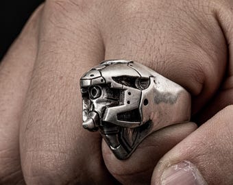 May include: A silver ring with a detailed robot head design. The ring is worn on a person's finger.