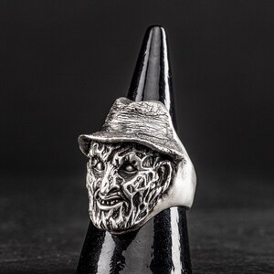 May include: A silver ring featuring a detailed, sculpted face with a hat. The face has a menacing expression with a wide, toothy grin.