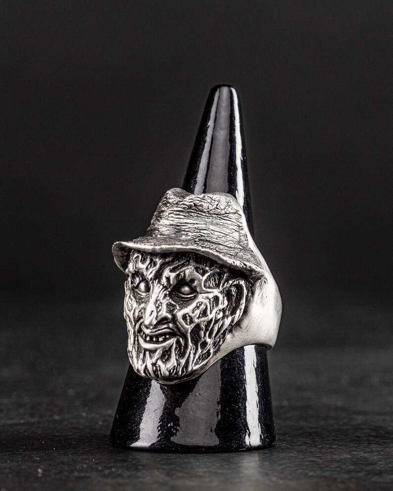 May include: A silver ring featuring a detailed, sculpted face with a hat. The face has a menacing expression with a wide, toothy grin.