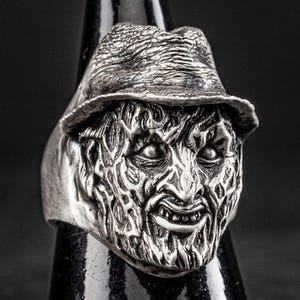 May include: A silver ring featuring a detailed, three-dimensional sculpture of a horror movie character's face. The character is wearing a hat and has a menacing expression.