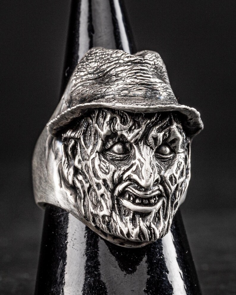 May include: A silver ring featuring a detailed, three-dimensional sculpture of a horror movie character's face. The character is wearing a hat and has a menacing expression.